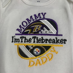 May include: White onesie with an embroidered design featuring a football helmet with the Baltimore Ravens logo and the text "Mommy I'm The Tiebreaker Daddy".
