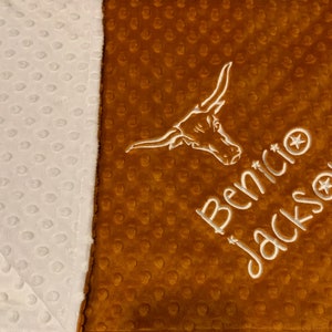 May include: A personalized baby blanket with a two-tone design. One side is white with a textured pattern, and the other is brown with a similar texture. The blanket features an embroidered longhorn logo and the name "Benicio Jackson" in white.