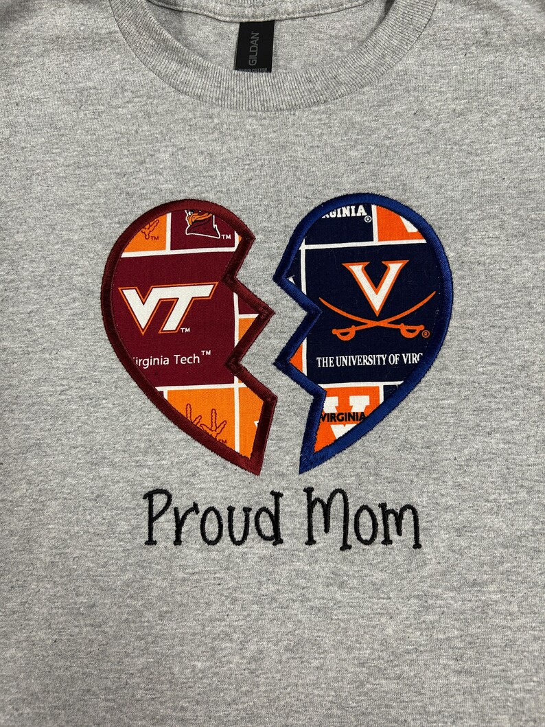 May include: A gray t-shirt featuring a broken heart design. One half displays the Virginia Tech logo in maroon, orange, and white. The other half shows the University of Virginia logo in navy and orange. The text "Proud Mom" is embroidered below.