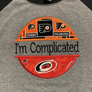 May include: A gray and black baseball t-shirt with a half-circle design featuring the Philadelphia Flyers logo and the Carolina Hurricanes logo. The text "I'm Complicated" is printed below the logos.