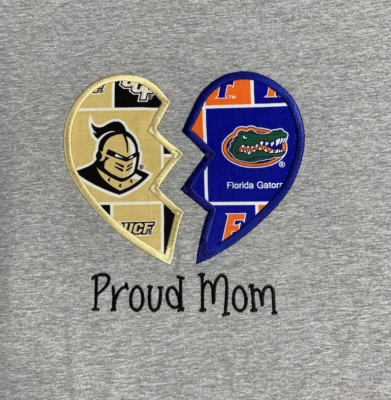 May include: A gray t-shirt featuring a broken heart design. One half displays the UCF Knights logo, the other the Florida Gators logo. Below the design, the words "Proud Mom" are embroidered in black.