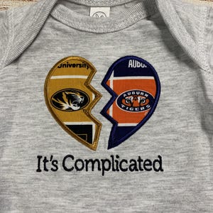 May include: A gray baby onesie with a broken heart design. One half features the University of Missouri logo, the other the Auburn Tigers logo. The phrase "It's Complicated" is embroidered below the heart.