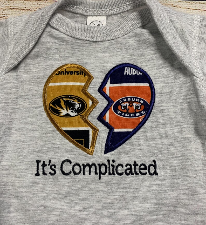 May include: A gray baby onesie with a broken heart design. One half features the University of Missouri logo, the other the Auburn Tigers logo. The phrase "It's Complicated" is embroidered below the heart.