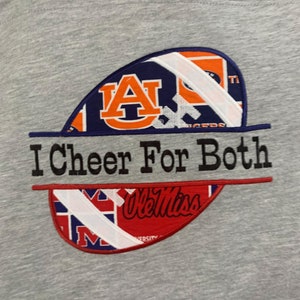 May include: Gray t-shirt with an embroidered design. The design features two football-shaped patches, one with the Auburn University logo and the other with the Ole Miss logo, separated by the text "I Cheer For Both".