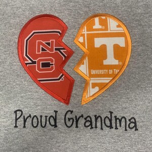 May include: A gray sweatshirt features a broken heart design with two halves. One half is red with "NC" and the other is orange with a "T" logo. The text "Proud Grandma" is embroidered below the heart.