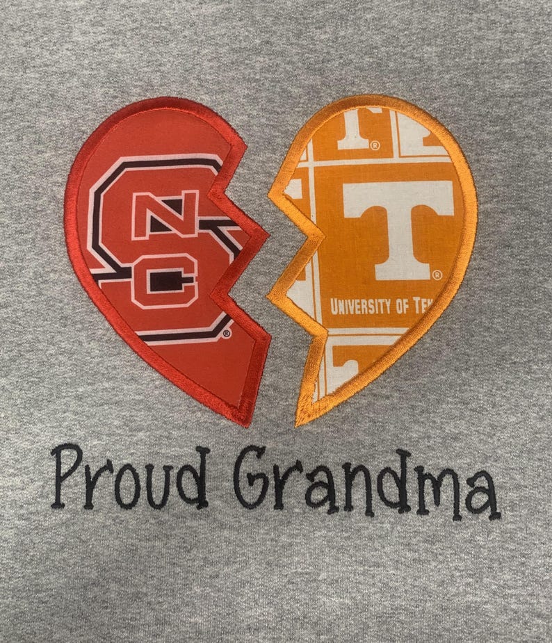 May include: A gray sweatshirt features a broken heart design with two halves. One half is red with "NC" and the other is orange with a "T" logo. The text "Proud Grandma" is embroidered below the heart.