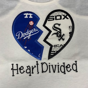 May include: White t-shirt featuring a split heart design. One half is blue with "Dodgers" and a baseball graphic, the other is black and white with "White Sox". Below the heart is the text "Heart Divided".