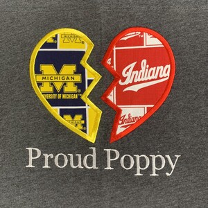 May include: Gray sweatshirt featuring a broken heart design. One half is yellow and navy blue with "Michigan" text, the other is red with "Indiana" text. The words "Proud Poppy" are embroidered below.