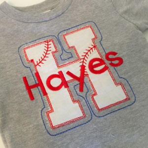 May include: A gray t-shirt with a white felt letter H with red stitching. The letter H has a baseball stitch design inside. The name "Hayes" is stitched in red below the letter.