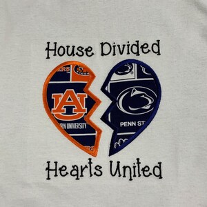 May include: White sweatshirt with a broken heart design. One half features the Auburn University logo in orange and navy, the other half displays the Penn State logo in navy and white. Text above reads "House Divided," below reads "Hearts United."