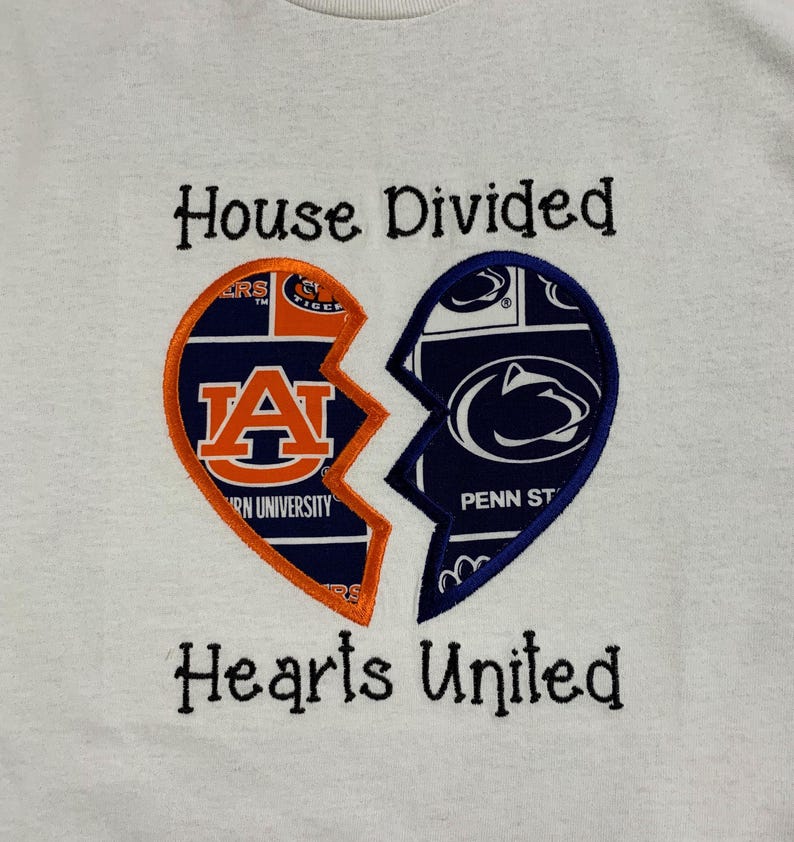 May include: White sweatshirt with a broken heart design. One half features the Auburn University logo in orange and navy, the other half displays the Penn State logo in navy and white. Text above reads "House Divided," below reads "Hearts United."