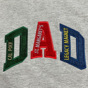 May include: A heather gray sweatshirt featuring the word DAD in large, block-style embroidered letters. The D is dark green with yellow text reading CAL POLY, the A is red with white text reading ST. MARGARET'S, and the D is blue with yellow text.
