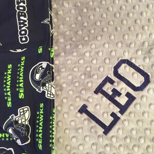 May include: A gray and white baby blanket with a blue embroidered name "LEO" on it. The blanket is made of a soft, textured fabric. The blanket is next to a blue fabric with a pattern of football helmets and the words "Seattle Seahawks" and "Cowboys".