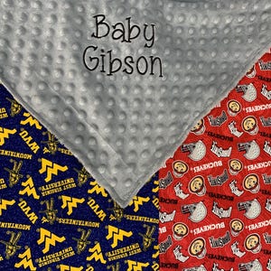 May include: A gray and white baby blanket with the text "Baby Gibson" embroidered on it. One side of the blanket features a blue and gold pattern with the West Virginia University Mountaineers logo. The other side features a red pattern with the Ohio State Buckeyes logo.