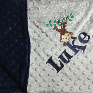 May include: A gray and navy blue baby blanket with a white and brown embroidered monkey hanging from a vine. The blanket has the name "Luke" embroidered on it.