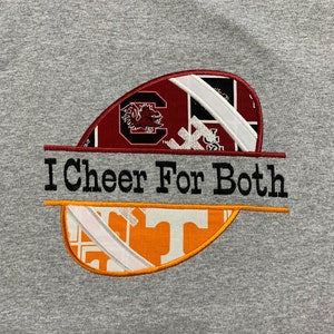 May include: Gray t-shirt with an embroidered football design. The football is made up of patches from the University of South Carolina and the University of Tennessee. The text "I Cheer For Both" is embroidered across the football.