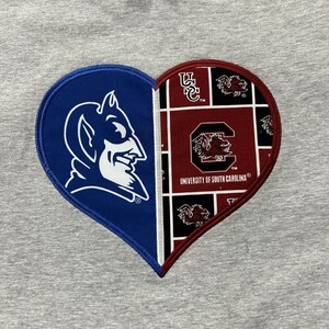 May include: A gray t-shirt featuring a heart-shaped design split into two halves. One side displays a blue devil mascot, the other a maroon and black pattern with the words "University of South Carolina".