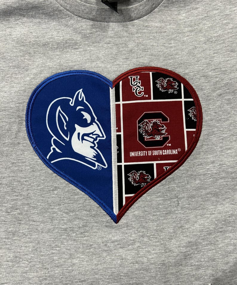 May include: A gray t-shirt featuring a heart-shaped design split into two halves. One side displays a blue devil mascot, the other a maroon and black pattern with the words "University of South Carolina".