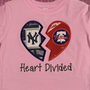 May include: Pink t-shirt featuring a broken heart design. One half displays the New York Yankees logo, the other the Philadelphia Phillies logo. The text "Heart Divided" is embroidered below the heart.