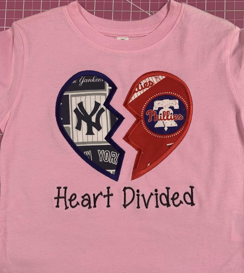 May include: Pink t-shirt featuring a broken heart design. One half displays the New York Yankees logo, the other the Philadelphia Phillies logo. The text "Heart Divided" is embroidered below the heart.