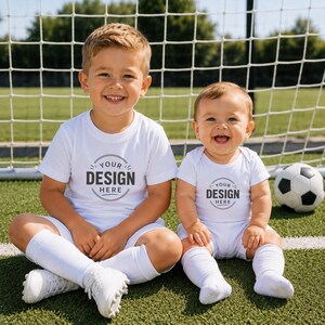 Puede incluir: Una camiseta blanca de manga corta y un body blanco de manga corta, ambos con un gr&aacute;fico circular con el texto "YOUR DESIGN HERE" en gris oscuro. Las prendas se llevan con calcetines deportivos blancos y botas de f&uacute;tbol blancas.
