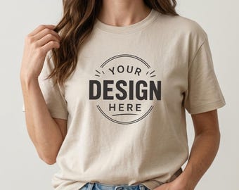 May include: A beige crew neck t-shirt featuring a black circular graphic on the chest with the text YOUR DESIGN HERE centered inside. The shirt has short sleeves and a standard fit, constructed from a solid-colored fabric with a smooth texture.