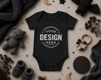 May include: A black short-sleeved baby bodysuit featuring the text YOUR DESIGN HERE inside a circular graphic. The garment has a round neckline and three snaps at the bottom. It is surrounded by a dark gray knit blanket, a plush bear, a bat, and wooden toys.