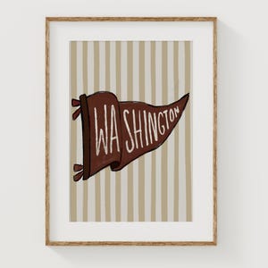 May include: A framed wall art print featuring a brown pennant with the white text WASHINGTON displayed across the centre. The pennant is set against a background of vertical beige and white stripes, all enclosed in a light brown wooden frame.