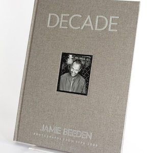 May include: A grey book with the title "DECADE" embossed in silver lettering. The book features a black and white photograph of a man on the front cover. The text "JAMIE BEEDEN PHOTOGRAPHS FROM 1998-2008" is printed in silver lettering below the photograph.