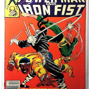 May include: Comic book cover for "Power Man and Iron Fist" with dynamic illustrations of the characters in battle. The cover is mainly orange, with the title in bold white and purple text. The text "Try fighting what can't be fought... Just try!" is also visible.
