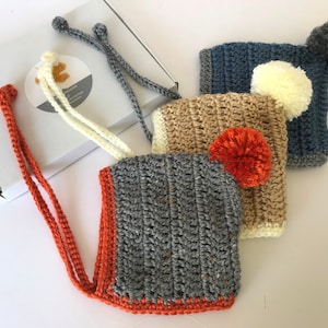May include: Three crocheted hats with pom-poms. The hats are in shades of grey, beige, and blue. Each hat has a different coloured pom-pom.