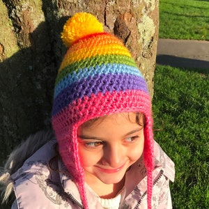 Peut inclure: Un bonnet en crochet rayé arc-en-ciel avec une bordure rose et un pompon jaune sur le dessus. Le bonnet a des rabats d'oreilles qui se nouent sous le menton.