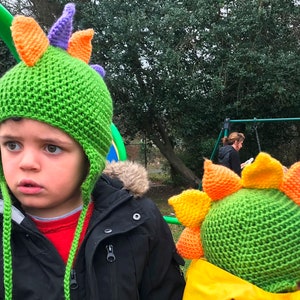 Peut inclure: Deux enfants portant des bonnets de dinosaure verts et jaunes avec des pointes. Les bonnets sont en crochet et ont des rabats d'oreilles.