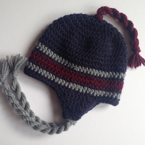 May include: A navy blue crocheted hat with ear flaps and a braided tassel. The hat features a horizontal stripe design in gray and burgundy. A gray braided cord extends from the ear flap. The hat is handmade.