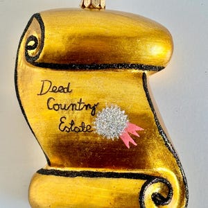 May include: A gold-colored Christmas ornament shaped like a scroll, with the words "Deed Country Estate" written on it. The scroll has black glitter trim and a silver and pink embellishment. A gold metal loop is at the top for hanging.