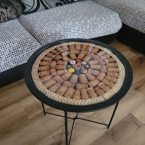 Op de afbeelding: Een ronde, zwarte metalen bijzettafel met een glazen blad. Het tafelblad is versierd met wijnkurken en bierdoppen. De tafel heeft een gevlochten rand en drie zwarte metalen poten. De tafel staat naast een bank met een patroon.