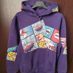 Può includere: Felpa con cappuccio viola con grafica in stile fumetto. Il design presenta fumetti con parole come "WOW!" e "BANG!" insieme a immagini di donne. La felpa ha una tasca frontale e un cappuccio con coulisse.