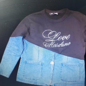 Può includere: Un top nero a maniche lunghe con la scritta "Love Moschino" in strass argentati. La metà inferiore è una giacca di jeans blu chiaro con due tasche frontali e un solo bottone bianco.