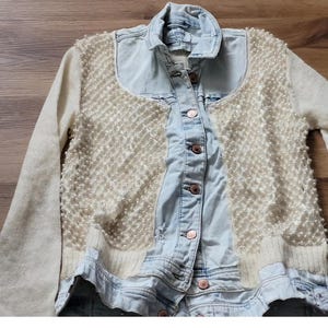 Puede incluir: Chaqueta vaquera azul claro con un panel frontal crema adornado con perlas. La chaqueta tiene cuello, mangas largas y botones color cobre. La palabra "Anteprima" es visible en la parte superior.