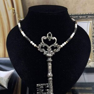 May include: A silver key pendant necklace with a pearl and bead chain. The key has an ornate design and is displayed on a black mannequin. The necklace is a vintage-inspired accessory.
