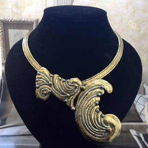 May include: A gold-toned statement necklace with an ornate, swirling design. The necklace features a wide, textured collar and a large, asymmetrical pendant with silver and gold accents. The jewelry is displayed on a black mannequin.