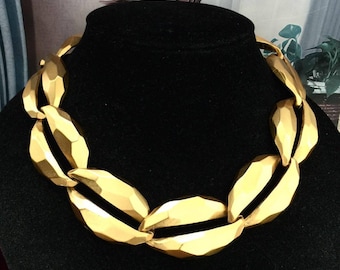 May include: A gold-toned statement necklace with a bold, geometric design. The necklace features large, faceted links that create a chunky, eye-catching look. The necklace is displayed on a black bust, highlighting its color and texture.