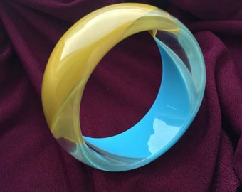 May include: A vibrant, two-tone bangle bracelet. The top half is a glossy yellow, transitioning to a bright blue on the bottom half. The bracelet is made of a smooth, possibly resin material, and is set against a deep purple fabric background.