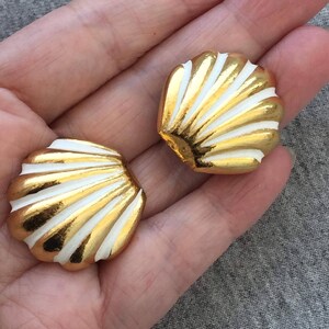 May include: A pair of seashell-shaped earrings. Each earring features a gold-colored outer edge and white, fan-like stripes. The earrings are designed to resemble a scallop shell.