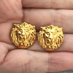 May include: Two gold-colored lion head studs. The studs are highly detailed, with a textured mane and facial features. The lion heads are facing forward, and the overall design is symmetrical.