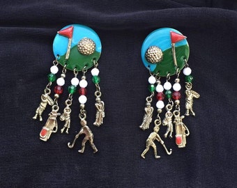 May include: A pair of golf-themed earrings. Each earring features a blue and green disc with a golf ball and flag. Dangling from the disc are beaded strands with golf player charms and golf bag charms. The earrings have red, white, and green beads.