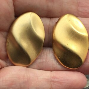 May include: A pair of gold-coloured, oval-shaped earrings with a textured, rippled surface. The earrings are held in a person's hand. The earrings have a matte finish and a slight curve.