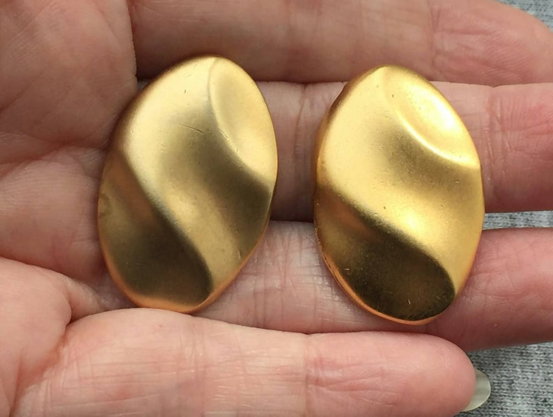 May include: A pair of gold-coloured, oval-shaped earrings with a textured, rippled surface. The earrings are held in a person's hand. The earrings have a matte finish and a slight curve.