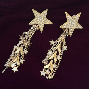 Puede incluir: Un par de pendientes de estrella dorados. Cada pendiente presenta una estrella grande incrustada con pedrería transparente. Cadenas de pedrería y pequeños dijes en forma de estrella cuelgan de las estrellas, creando un efecto de cascada. El fondo es de color burdeos oscuro.