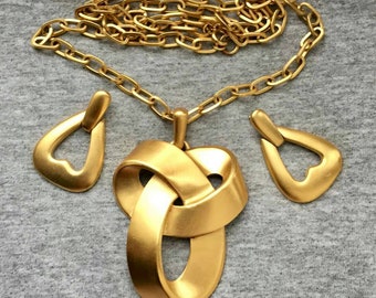 Puede incluir: Un conjunto de joyas de color dorado con un collar con un colgante grande e entrelazado y pendientes a juego en forma de lágrima. El collar tiene un diseño de eslabones de cadena. El conjunto se muestra sobre una superficie gris.